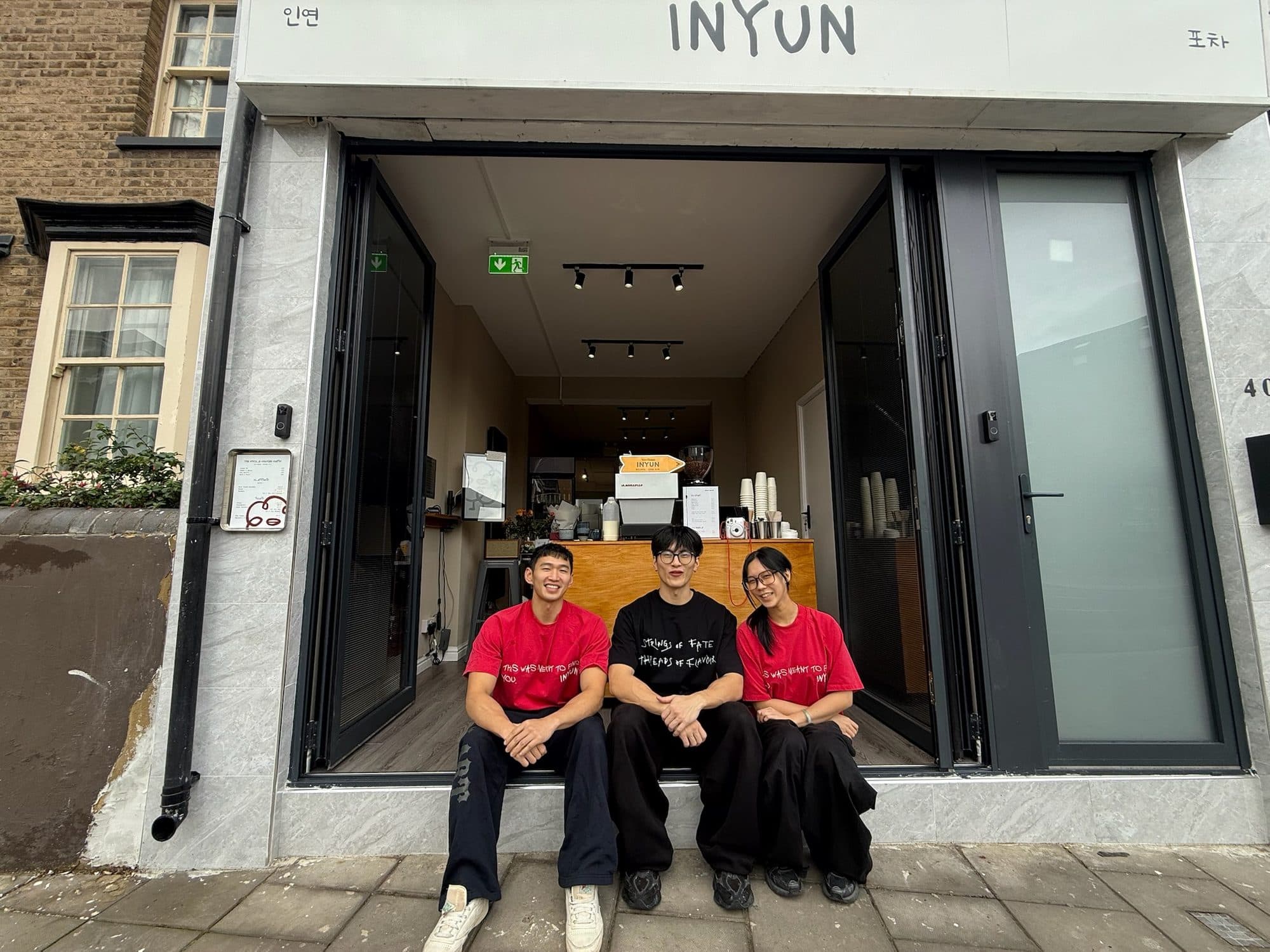 INYUN's three founders sitting together in the open shopfront doorway on Brentford High Street, under the hand-drawn sign